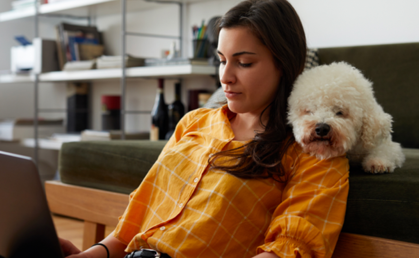 Woman sitting in front of couch while using laptop with a dog resting on her shoulder